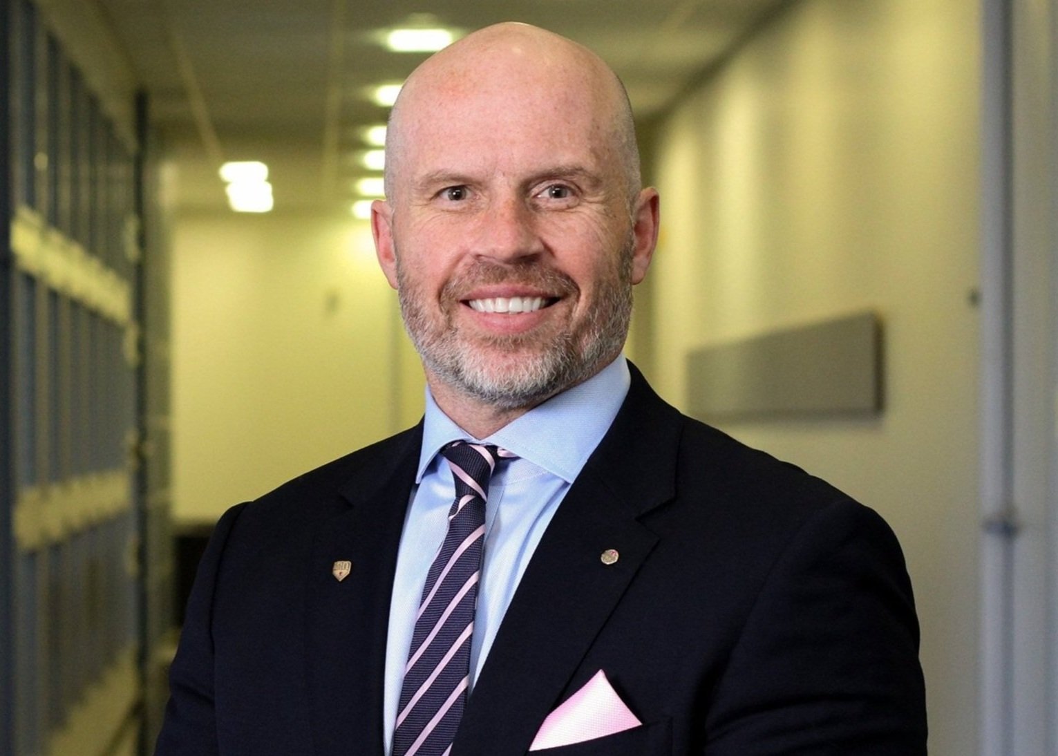 A middle-aged man with a bald head and a beard, smiling, dressed in a suit with a striped tie and a pink pocket square, standing in a hallway with lockers.