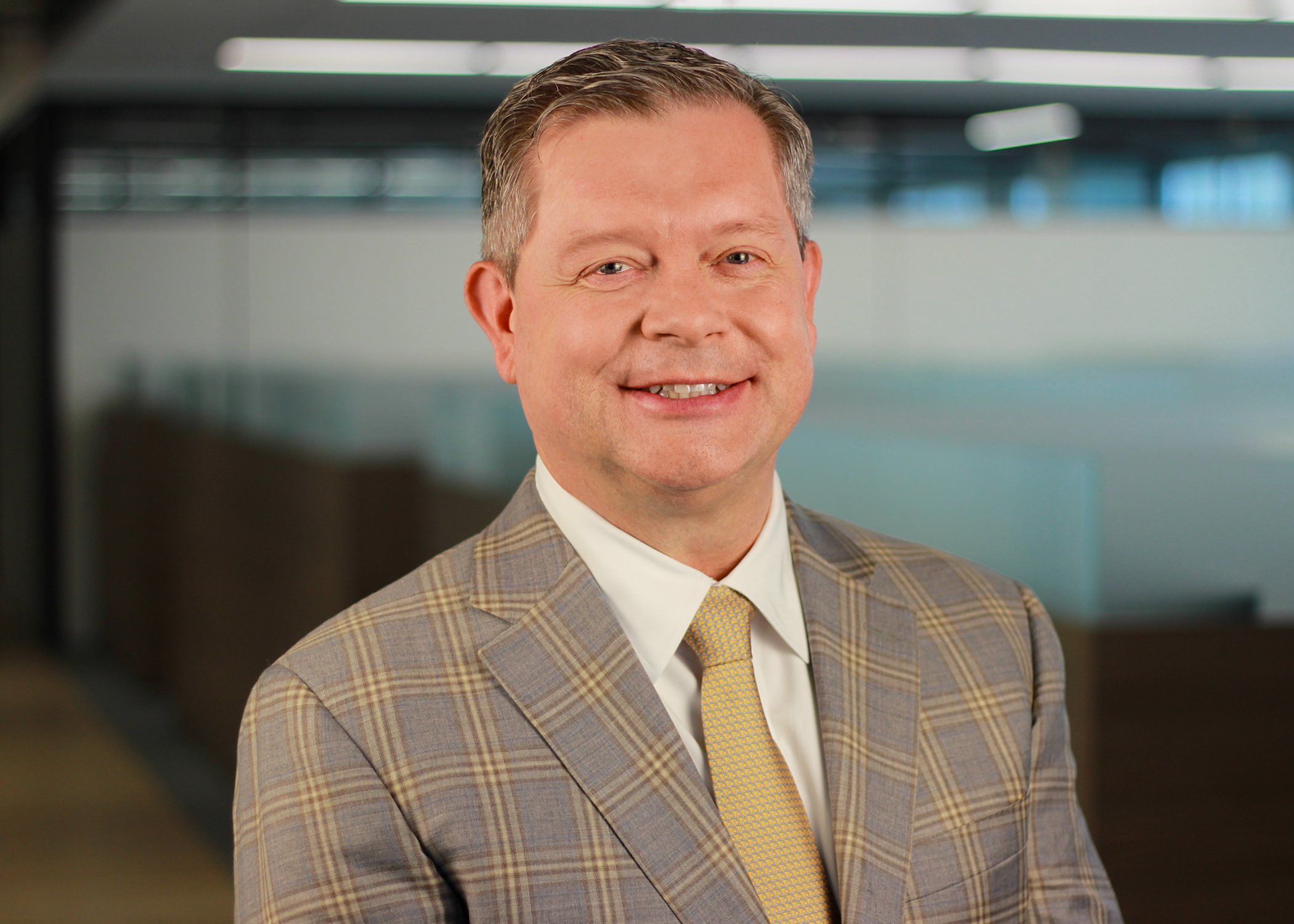 A middle-aged man in a checkered blazer and yellow tie smiling at the camera in an office setting.