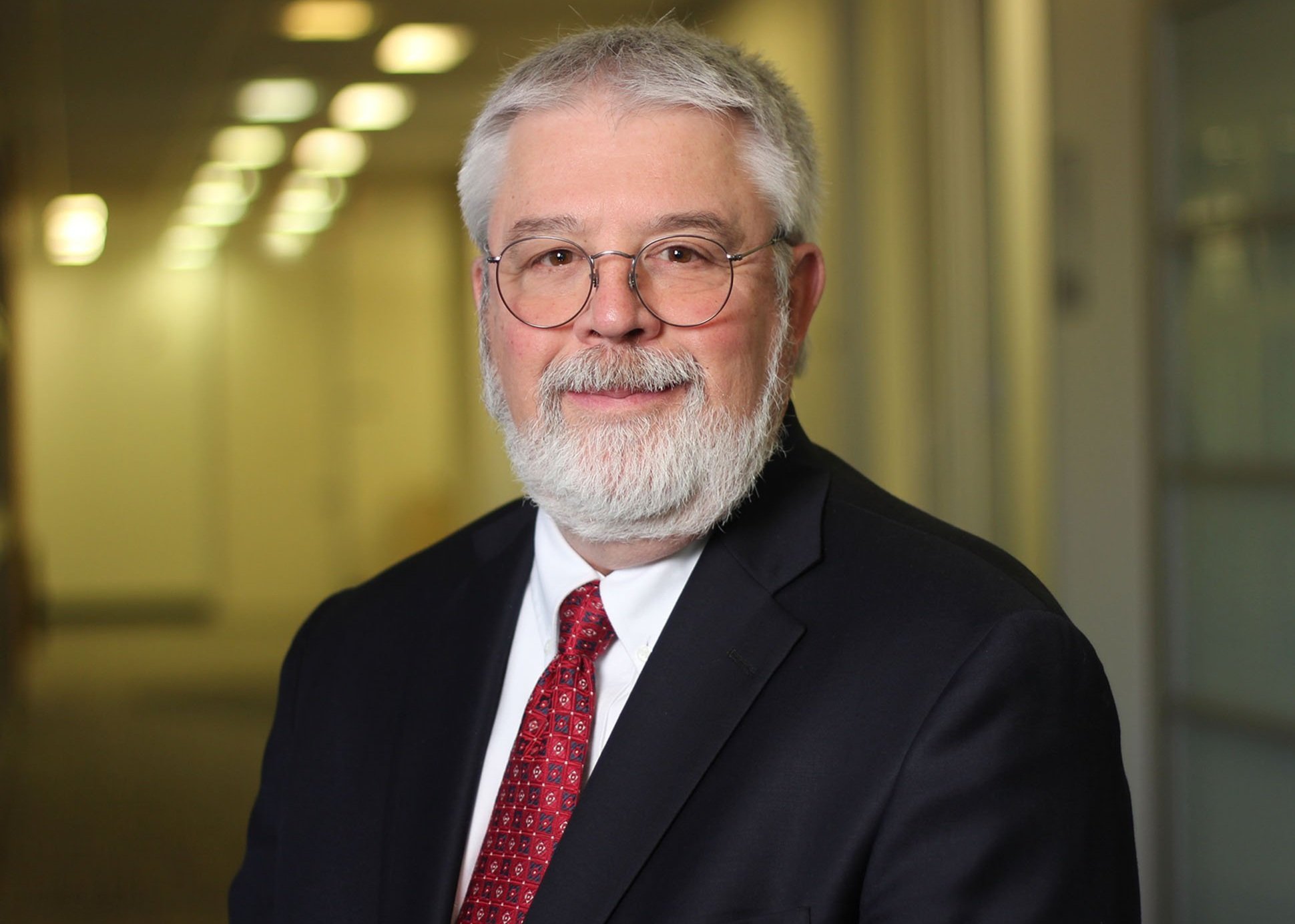 Portrait of an older man with white hair, a beard, glasses, wearing a black suit, white shirt, and red patterned tie, standing in a corridor with blurred lights and windows in the background.