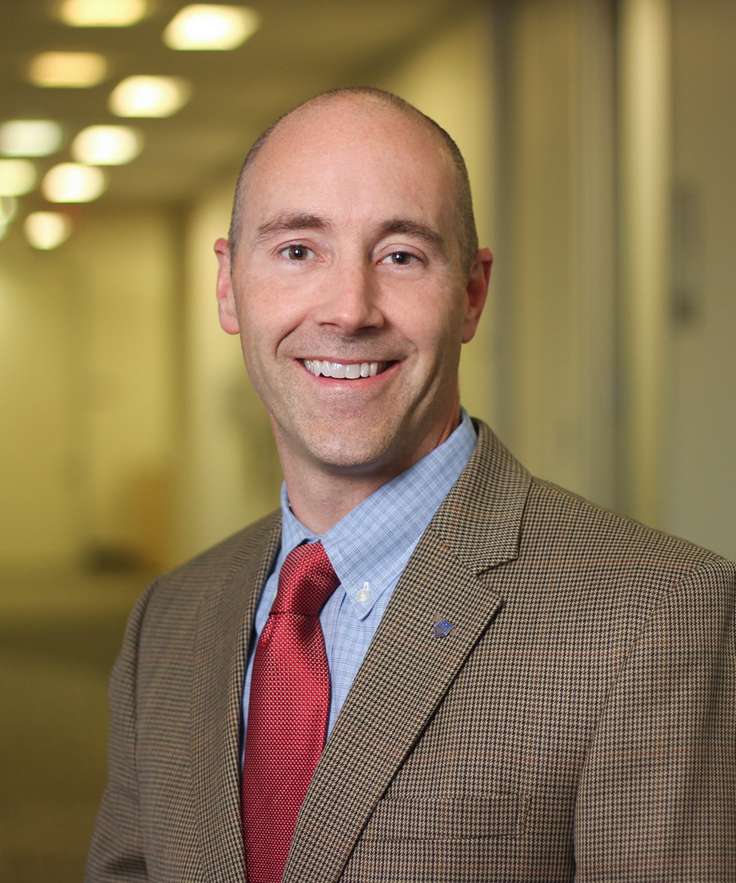 A smiling man in a brown checkered blazer, light blue shirt, and red tie standing in a well-lit hallway.