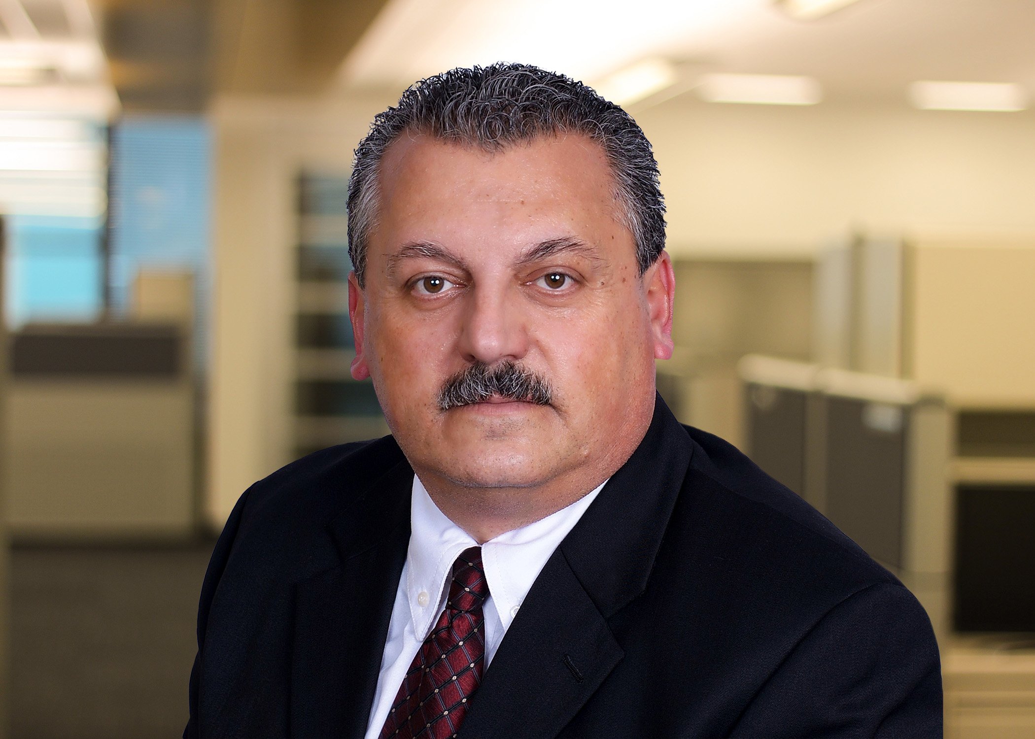 A middle-aged man with dark hair, a mustache, wearing a dark suit, white shirt, and maroon tie posed in an office setting.