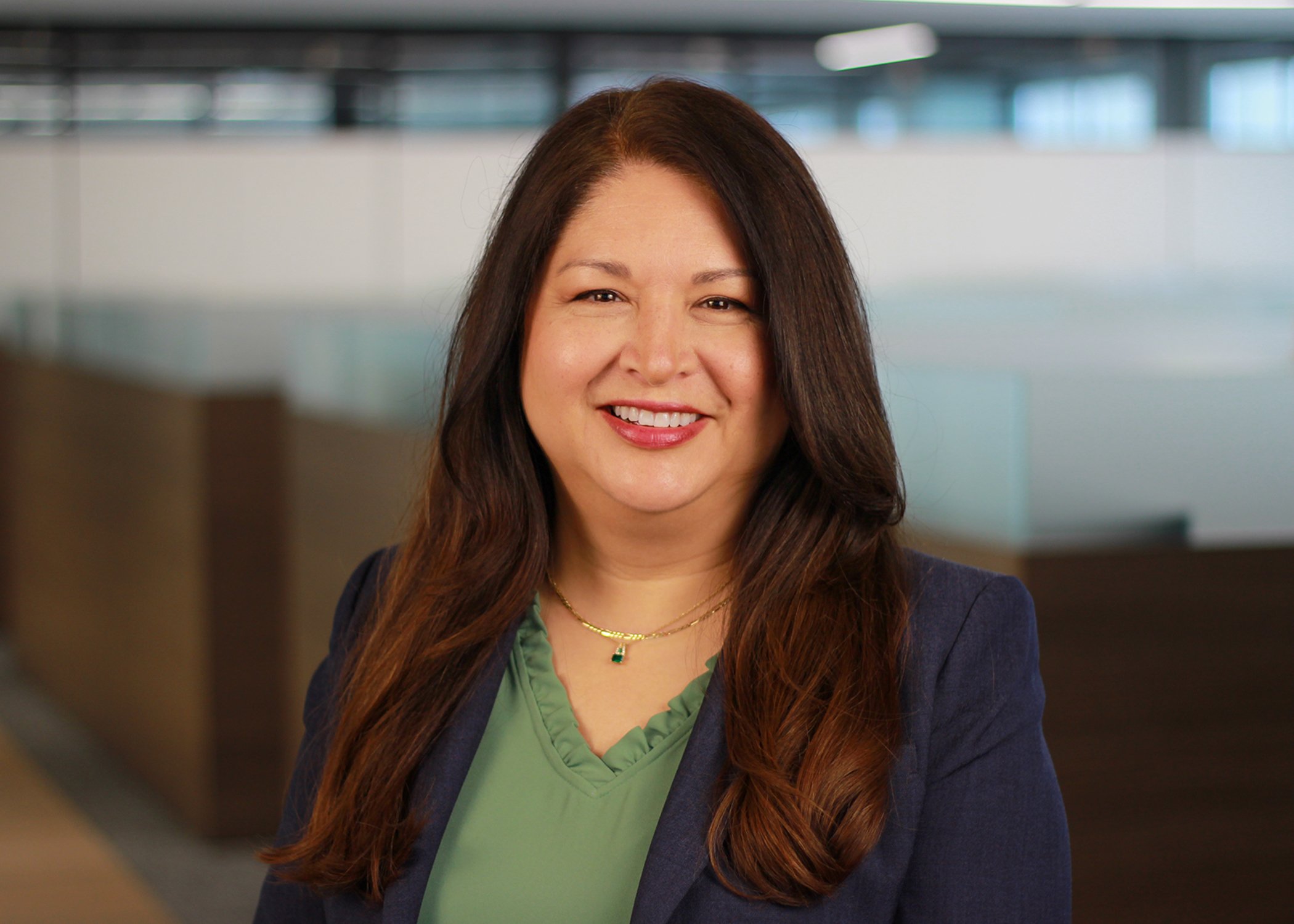 A woman with long brown hair, wearing a green top and a navy blazer, smiling in an indoor office setting.