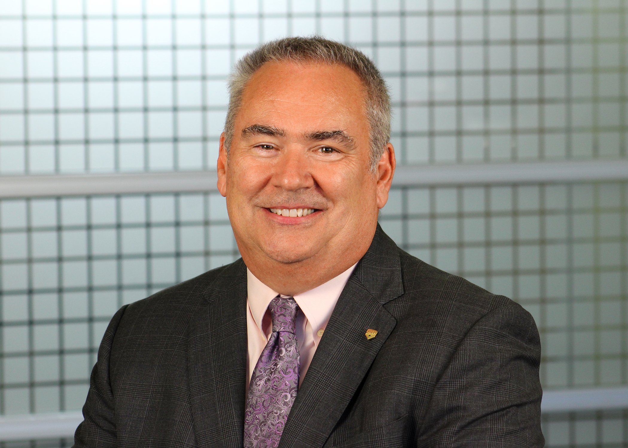 A middle-aged man with gray hair smiling in a professional suit and tie, standing in front of a modern glass building.